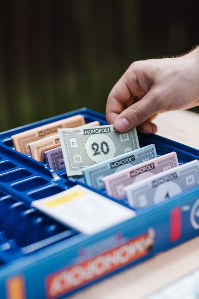 Crop unrecognizable person showing game banknote from collection in box while playing Monopoly at table